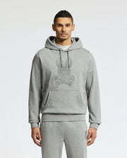 A person stands in a Psycho Bunny ELDORADO Brush Back Hoodie - B6H234E200, perfect for swaddle season. This cozy gray hoodie has a striking skull design, matching sweatpants, and features a large front pocket with drawstrings made from soft brush-back cotton. The background is plain white.