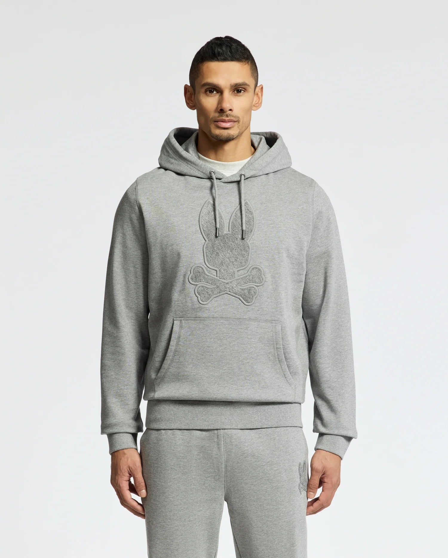 A person stands in a Psycho Bunny ELDORADO Brush Back Hoodie - B6H234E200, perfect for swaddle season. This cozy gray hoodie has a striking skull design, matching sweatpants, and features a large front pocket with drawstrings made from soft brush-back cotton. The background is plain white.