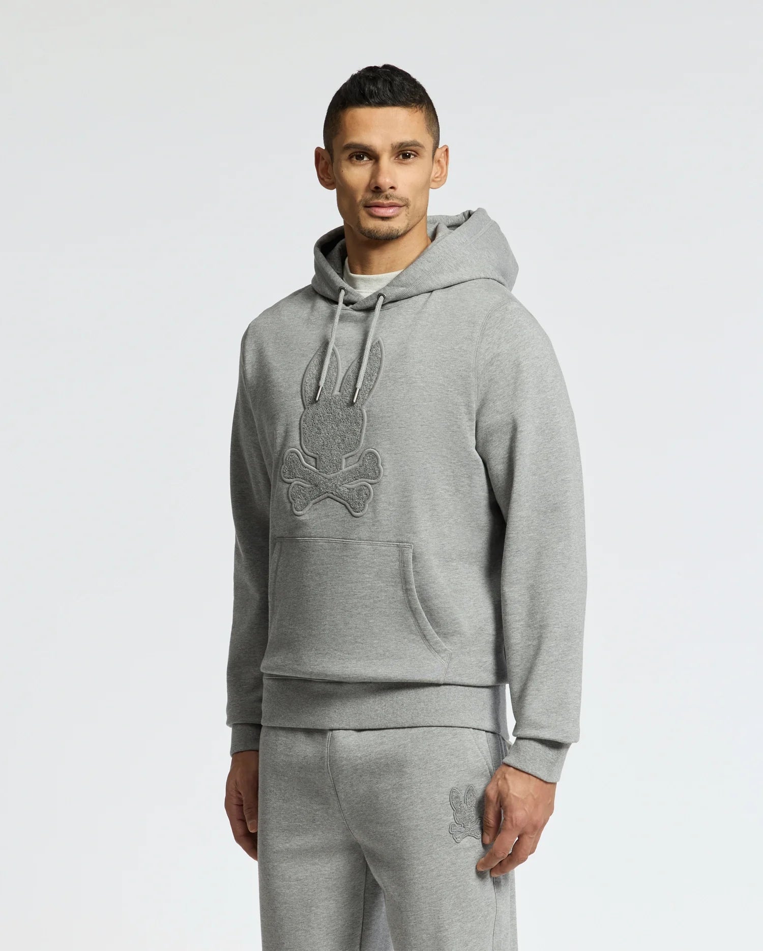 A man wears the Psycho Bunny MENS ELDORADO BRUSH BACK HOODIE, a light gray cotton hoodie with a dark gray bunny and crossbones emblem. He pairs it with matching sweatpants, posing against a plain white backdrop and maintaining a neutral expression.