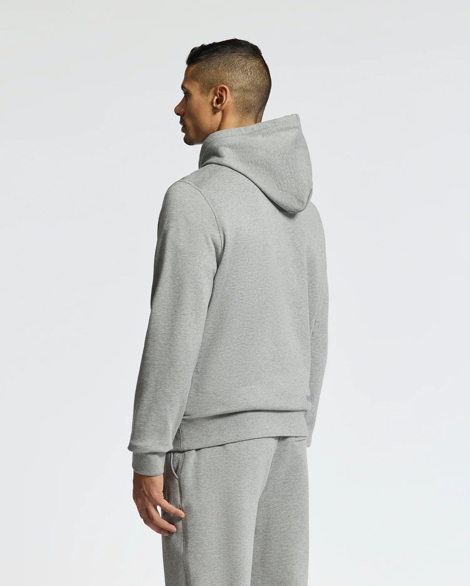 A person in the Psycho Bunny Men's Eldorado Brush Back Hoodie, B6H234E200, and matching sweatpants stands facing away. With the hood down, they glance left against a plain white backdrop—ideal attire for cozy days ahead in swaddle season.