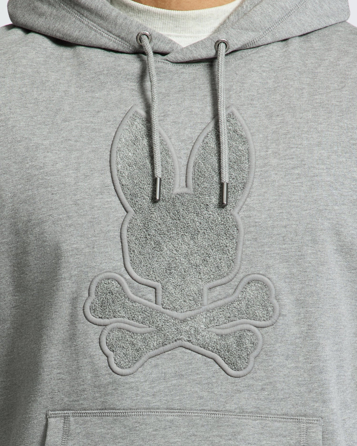 Close-up of the Psycho Bunny Men's Eldorado Brush Back Hoodie (B6H234E200) in gray, featuring a textured rabbit head with elongated ears above crossed bones, mimicking a skull and crossbones design. The motif is in a subtly darker shade—ideal for swaddle season.