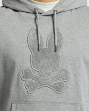 Close-up of the Psycho Bunny Men's Eldorado Brush Back Hoodie (B6H234E200) in gray, featuring a textured rabbit head with elongated ears above crossed bones, mimicking a skull and crossbones design. The motif is in a subtly darker shade—ideal for swaddle season.