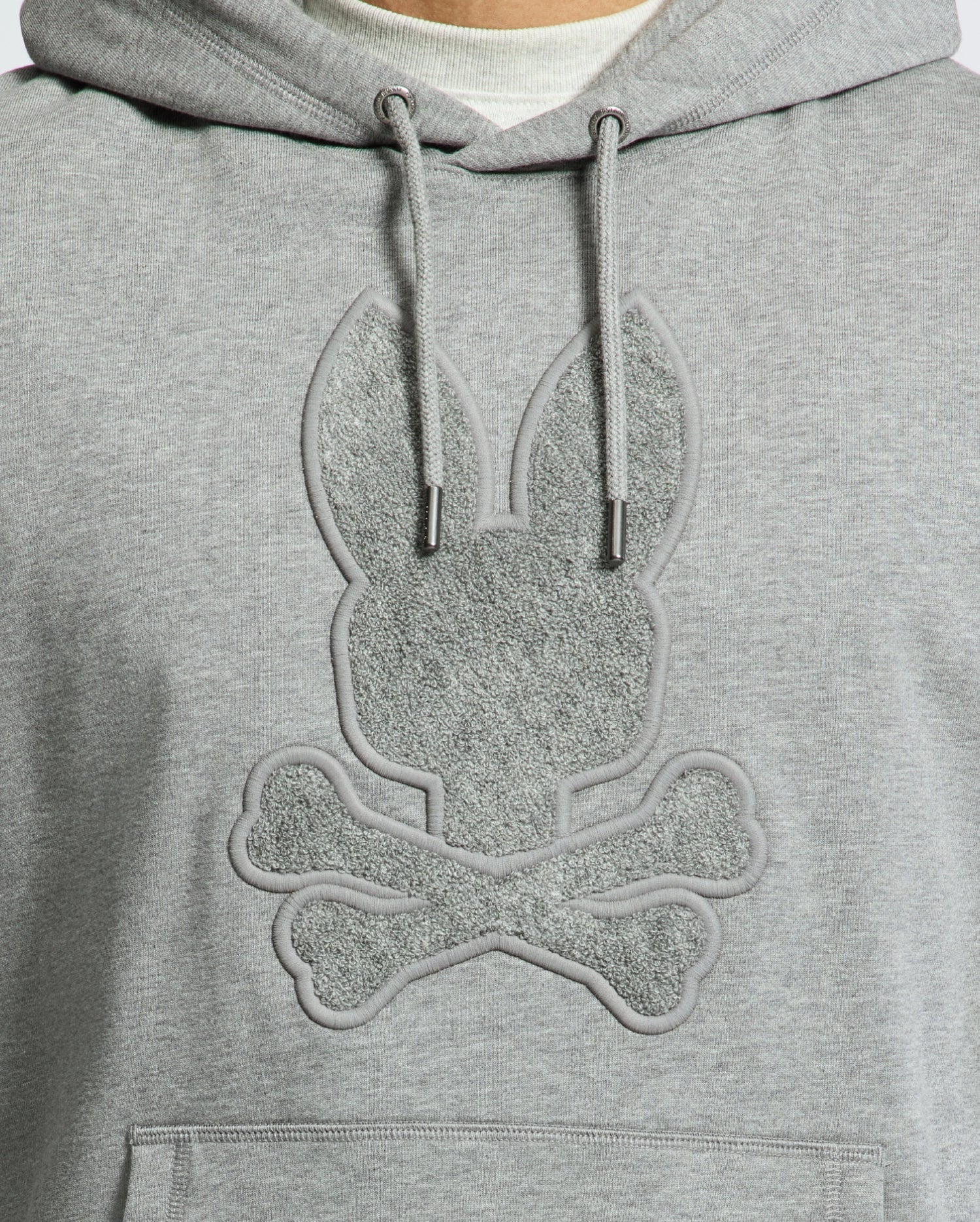 Close-up of the Psycho Bunny Men's Eldorado Brush Back Hoodie (B6H234E200) in gray, featuring a textured rabbit head with elongated ears above crossed bones, mimicking a skull and crossbones design. The motif is in a subtly darker shade—ideal for swaddle season.