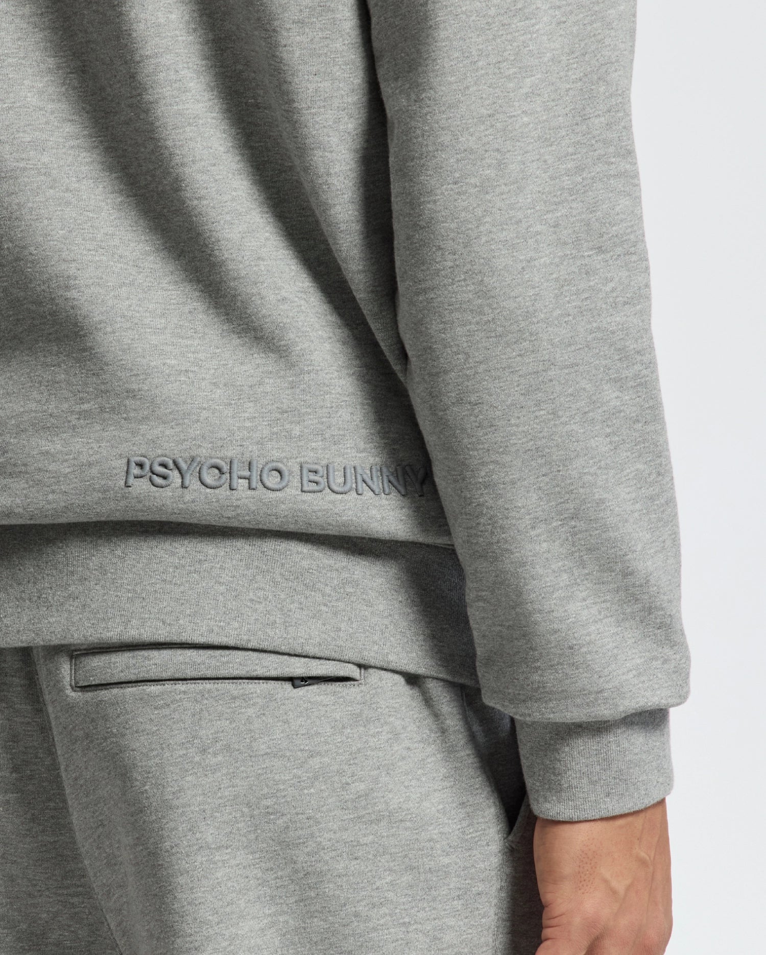 Dressed for swaddle season, a person wears the gray MENS ELDORADO BRUSH BACK HOODIE by Psycho Bunny, made from cozy brush-back cotton. Paired with matching gray pants, only their side and back are visible.