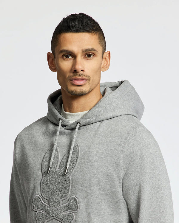 A person wears the Psycho Bunny MENS ELDORADO BRUSH BACK HOODIE (B6H234E200) in gray, featuring a textured skull with bunny ears. The cozy brush-back cotton makes it swaddle-season ready, and it’s styled with a drawstring hood against a light background.