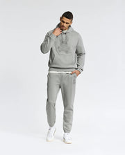Against a white backdrop, a person in Psycho Bunny's MENS ELDORADO Brush Back Cotton Hoodie and sweatpants lounges comfortably. Clad in white sneakers, their right hand grazes their face as they look down, embodying the cozy essence of swaddle season.