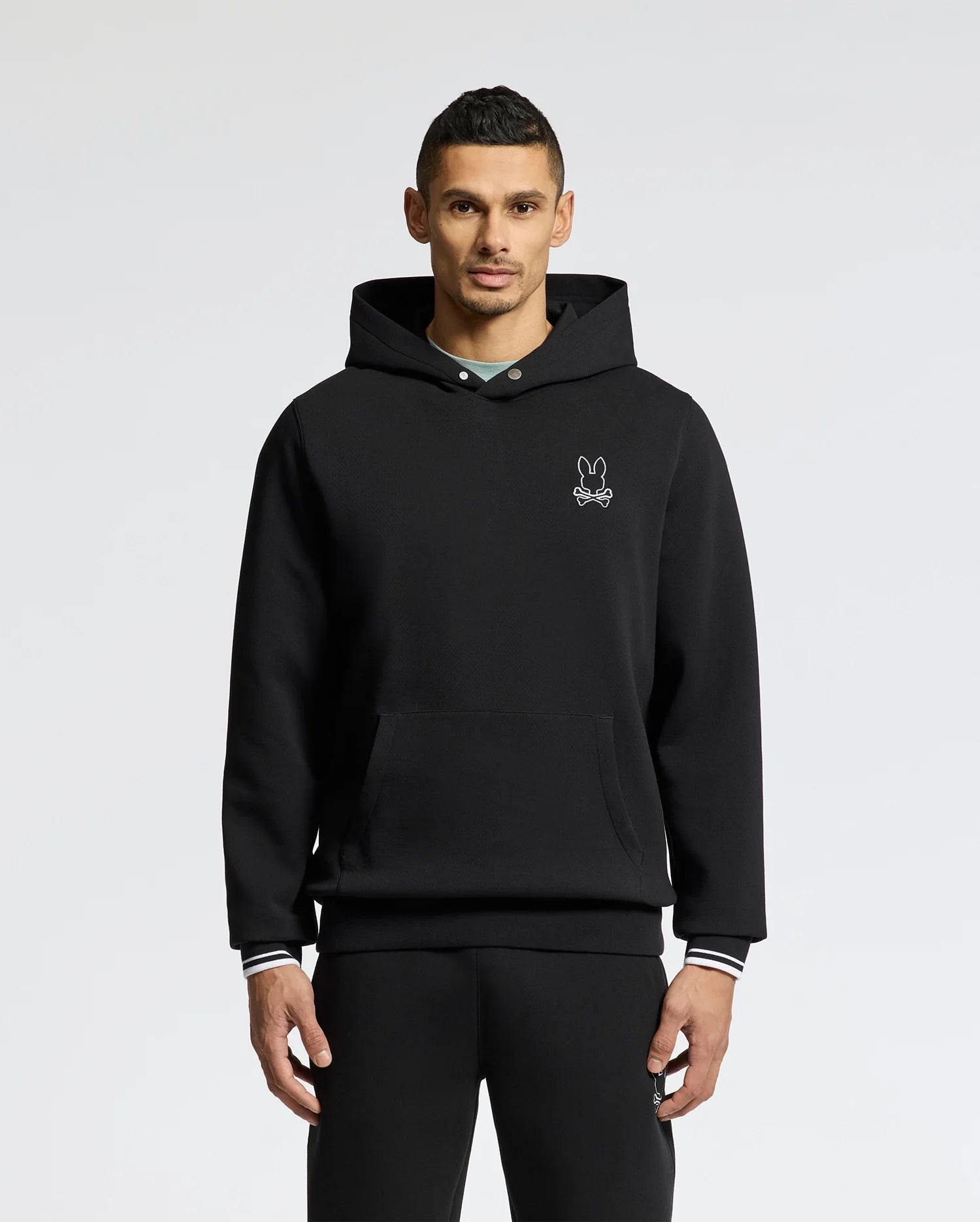 A person in a Psycho Bunny MENS DACANO PERFORATED HOODIE - B6H239E200, featuring a breathable design and embroidered bunny logo on the chest, stands against a plain white background. They have short dark hair and look directly at the camera with hands by their sides.