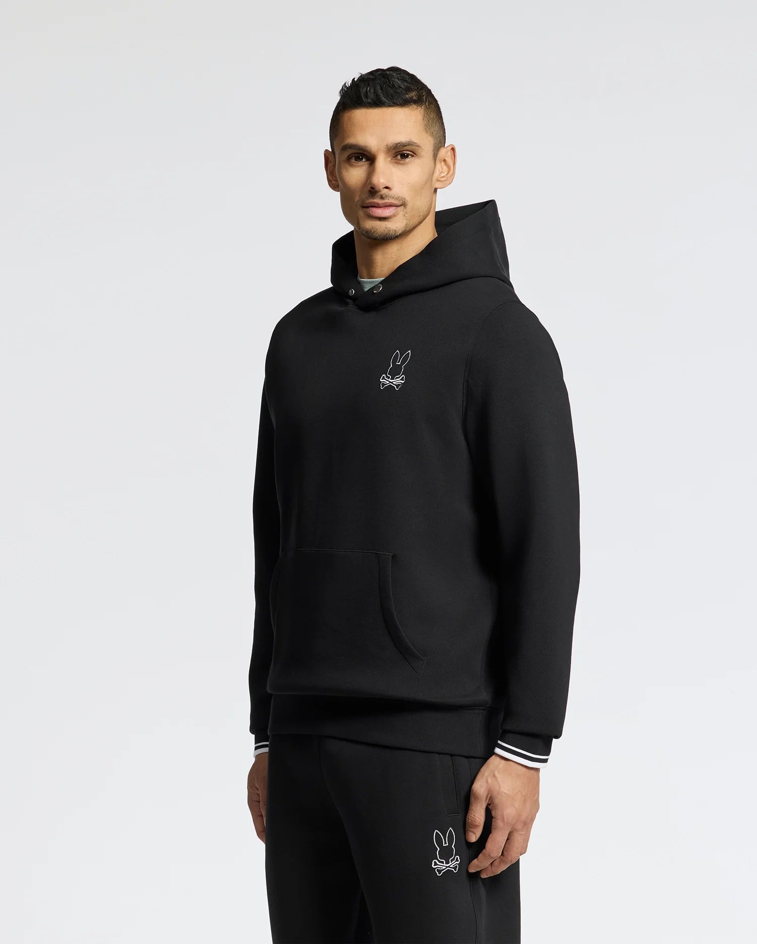 A person wears the Psycho Bunny MENS DACANO PERFORATED HOODIE - B6H239E200, a black breathable hoodie and pants with a small embroidered rabbit design. The hoodie features a pocket and white-striped cuffs. They stand against a plain white background, gazing into the camera.