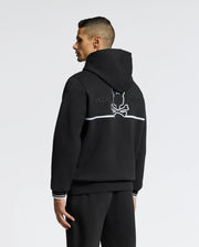 A person in the Psycho Bunny MENS DACANO PERFORATED HOODIE, black with white trim, faces left. The back features a meticulously embroidered "PSYCHO BUNNY" logo displaying a rabbit silhouette with crossbones beneath.
