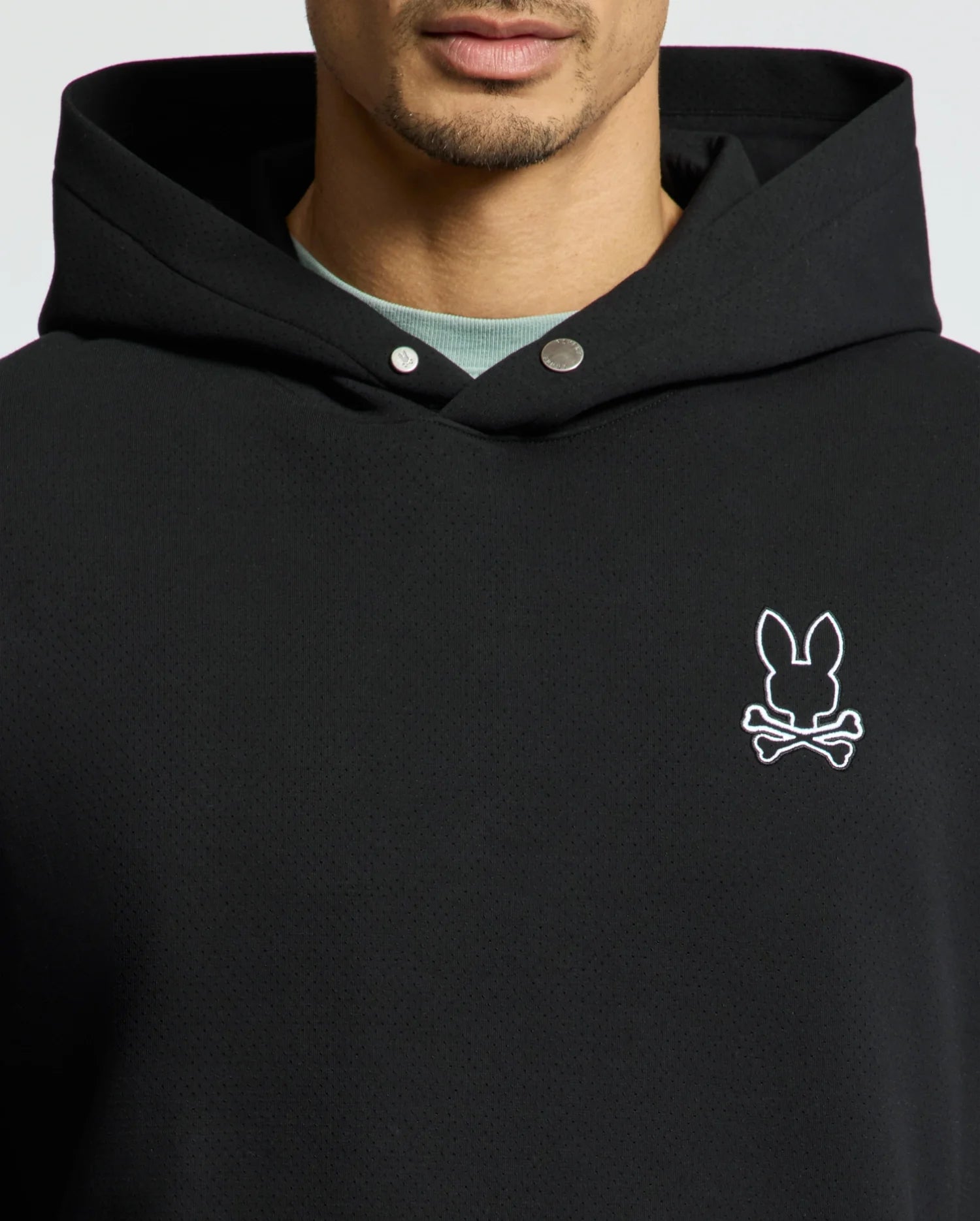 A person is wearing the Psycho Bunny MENS DACANO PERFORATED HOODIE - B6H239E200, a breathable black hoodie with an embroidered bunny and crossbones emblem on the chest and silver snap buttons near the neckline, partially revealing a light shirt underneath.