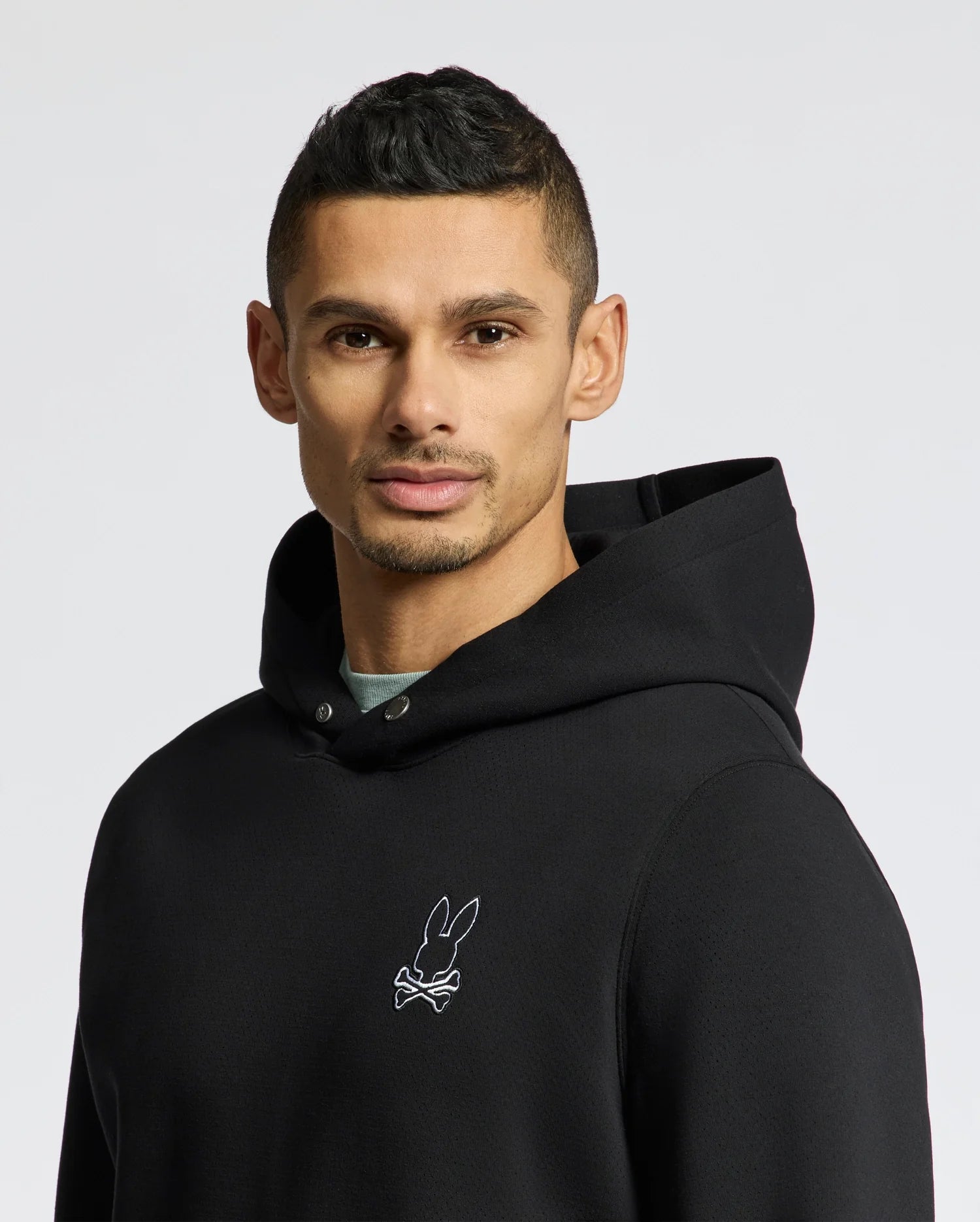 A person wearing the Psycho Bunny Men's Dacano Perforated Hoodie in black, featuring a breathable design and an embroidered logo of a rabbit head with crossbones on the chest, stands against a plain background.