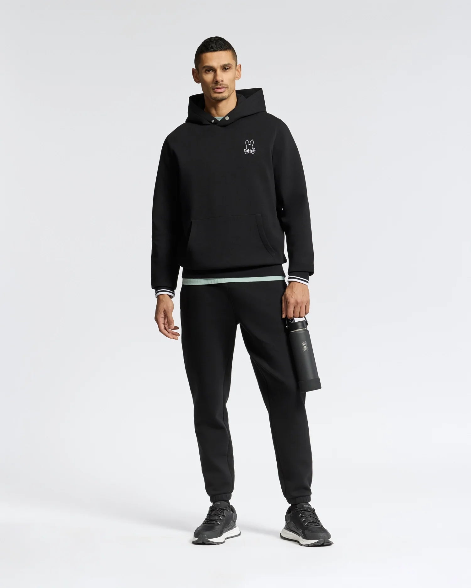 Wearing a Psycho Bunny MENS DACANO PERFORATED HOODIE in black with an embroidered logo, black joggers, and sleek black sneakers, the person stands holding a black water bottle against a plain white background.