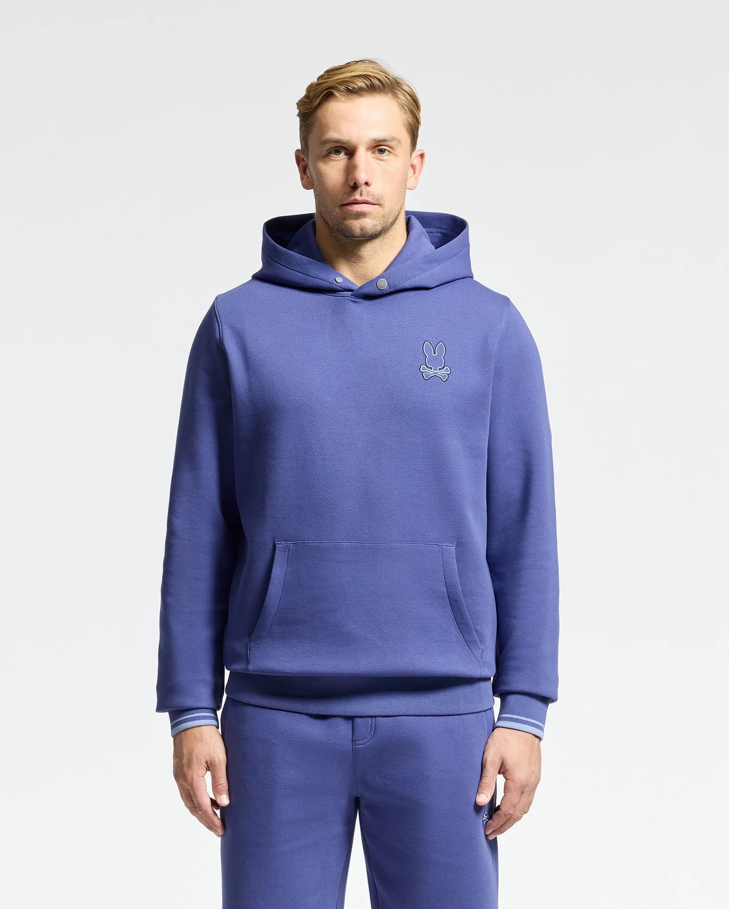 A person with short hair stands against a plain backdrop, wearing Psycho Bunny's DACANO Perforated Hoodie and matching sweatpants in blue stretch cotton-blend jersey. The hoodie displays a small embroidered rabbit logo on the left side, while their hands rest by their sides.