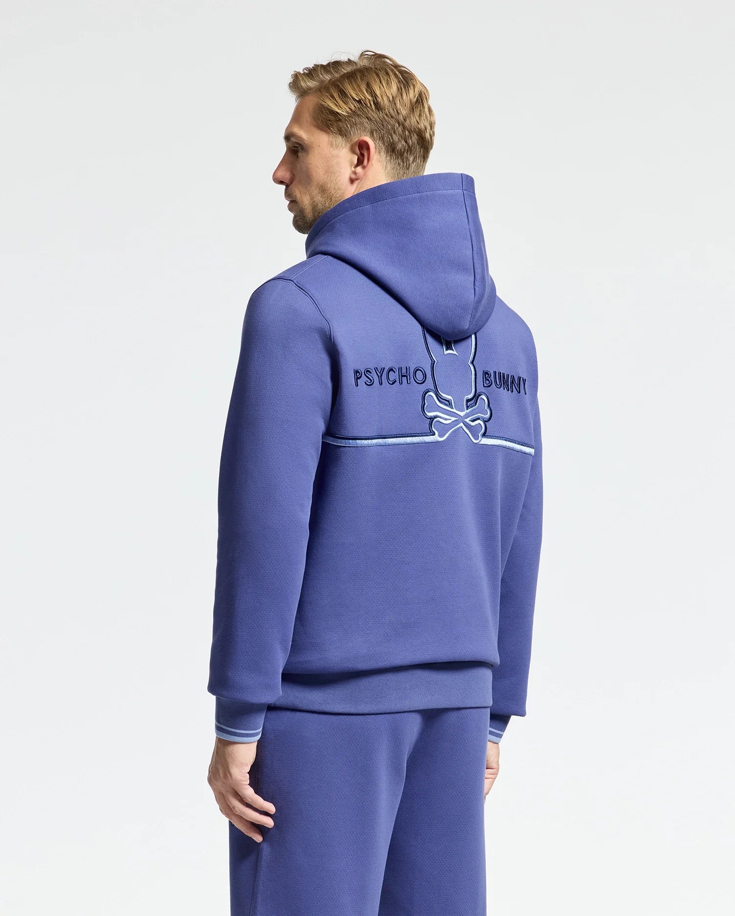 A person wearing the Psycho Bunny Mens Dacano Perforated Hoodie (B6H239E200) and matching pants stands against a white background; the hoodie back displays "Psycho Bunny" and an embroidered bunny and bones logo on comfortable stretch cotton-blend jersey.