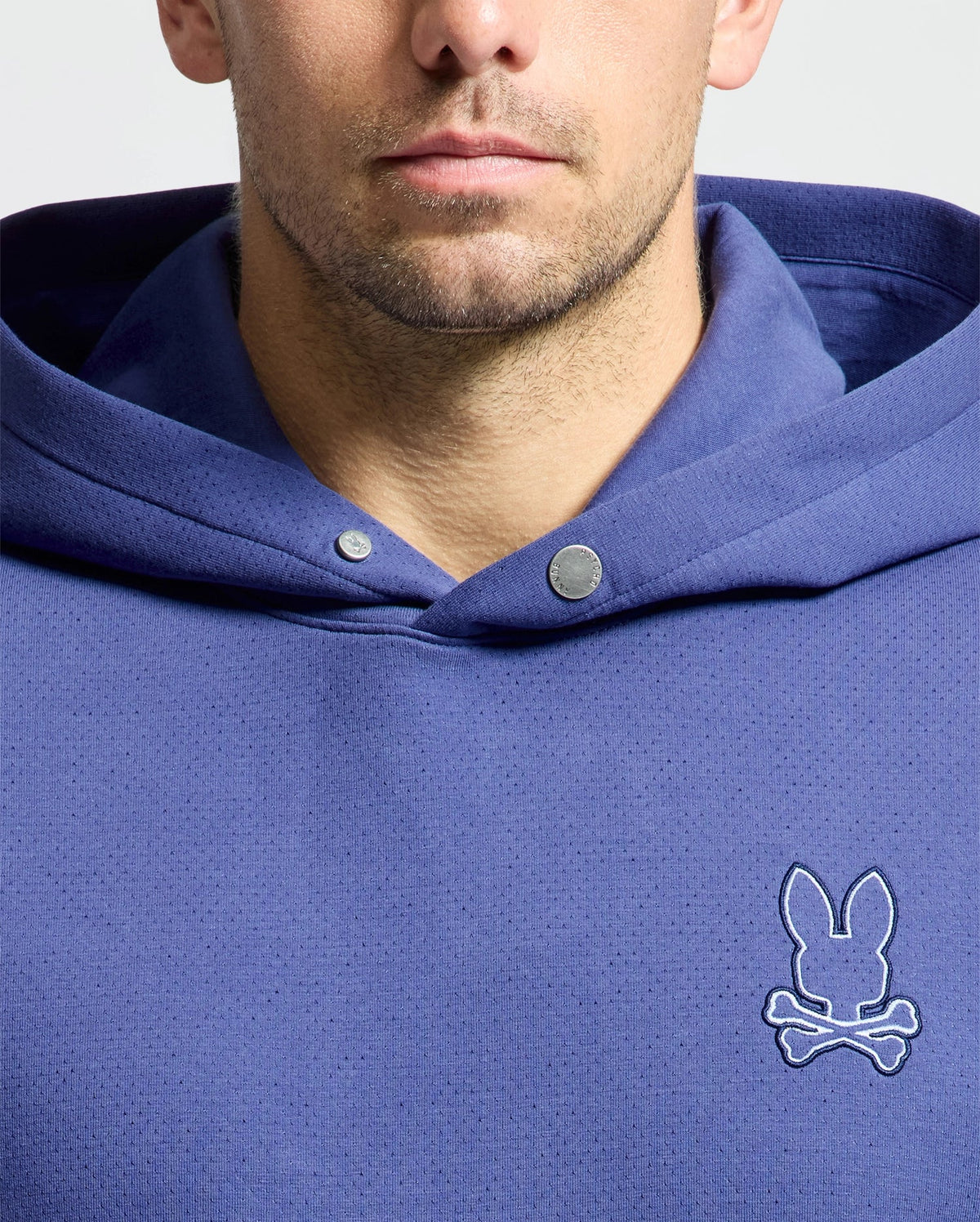 A man wears the Psycho Bunny MENS DACANO PERFORATED HOODIE (B6H239E200), a blue stretch cotton-blend jersey sweatshirt with snap buttons at the collar and an embroidered bunny over crossed bones logo on the chest. The image highlights the upper torso, fading near his chin.