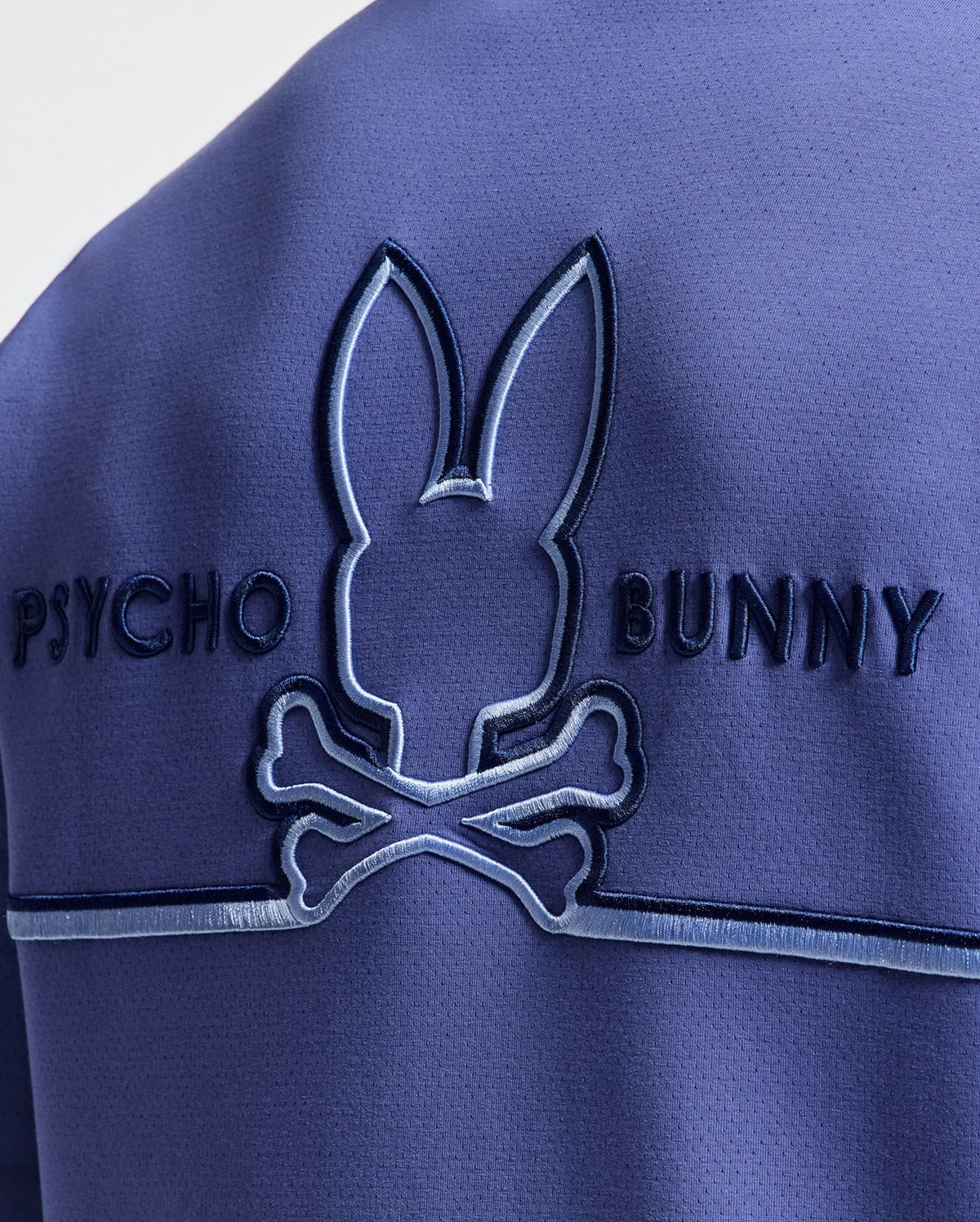 Close-up of a blue fabric on the Psycho Bunny MENS DACANO PERFORATED HOODIE with an embroidered bunny logo above crossed bones and "Psycho Bunny" embossed beside it for an edgy look.