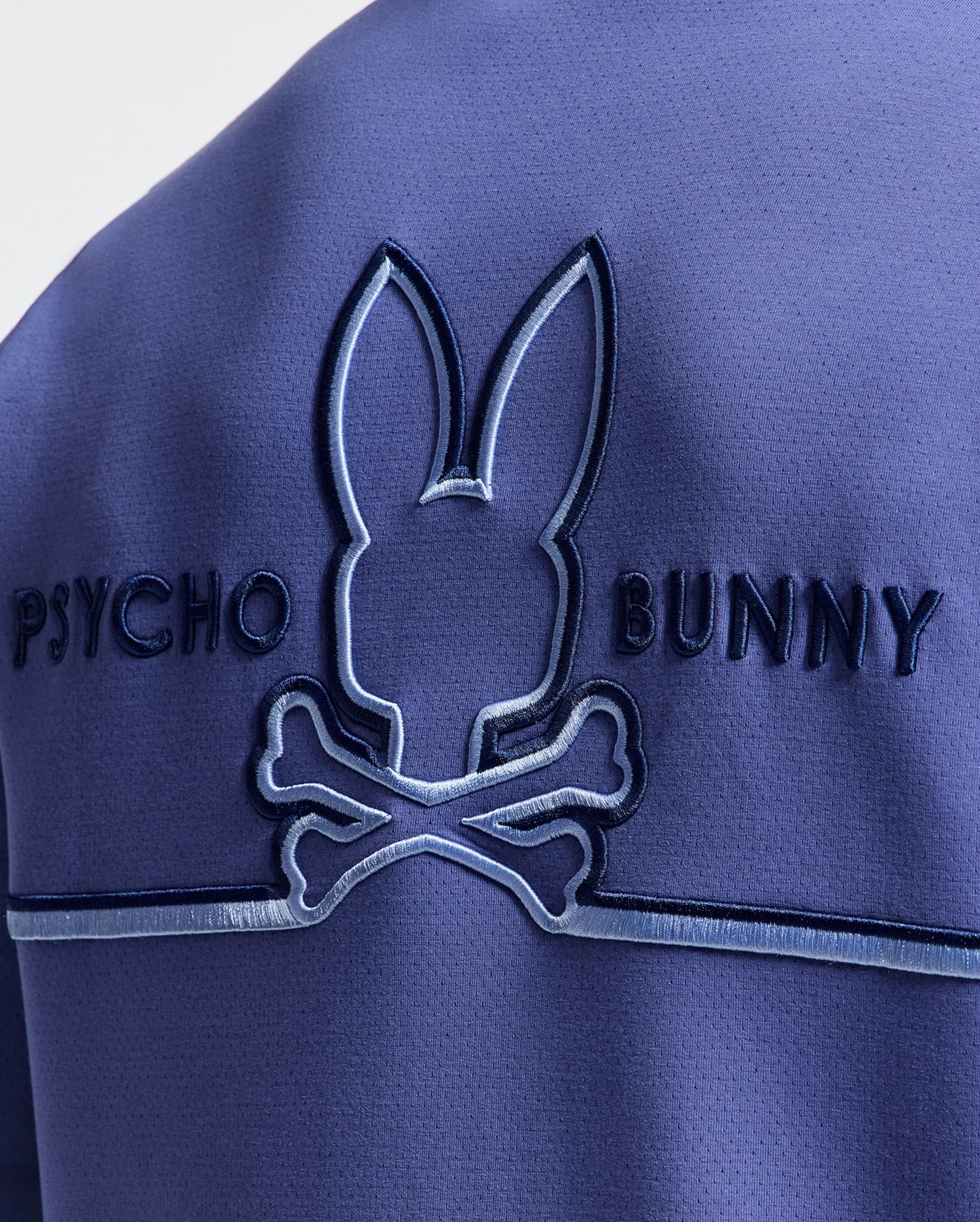 Close-up of a blue fabric on the Psycho Bunny MENS DACANO PERFORATED HOODIE with an embroidered bunny logo above crossed bones and "Psycho Bunny" embossed beside it for an edgy look.