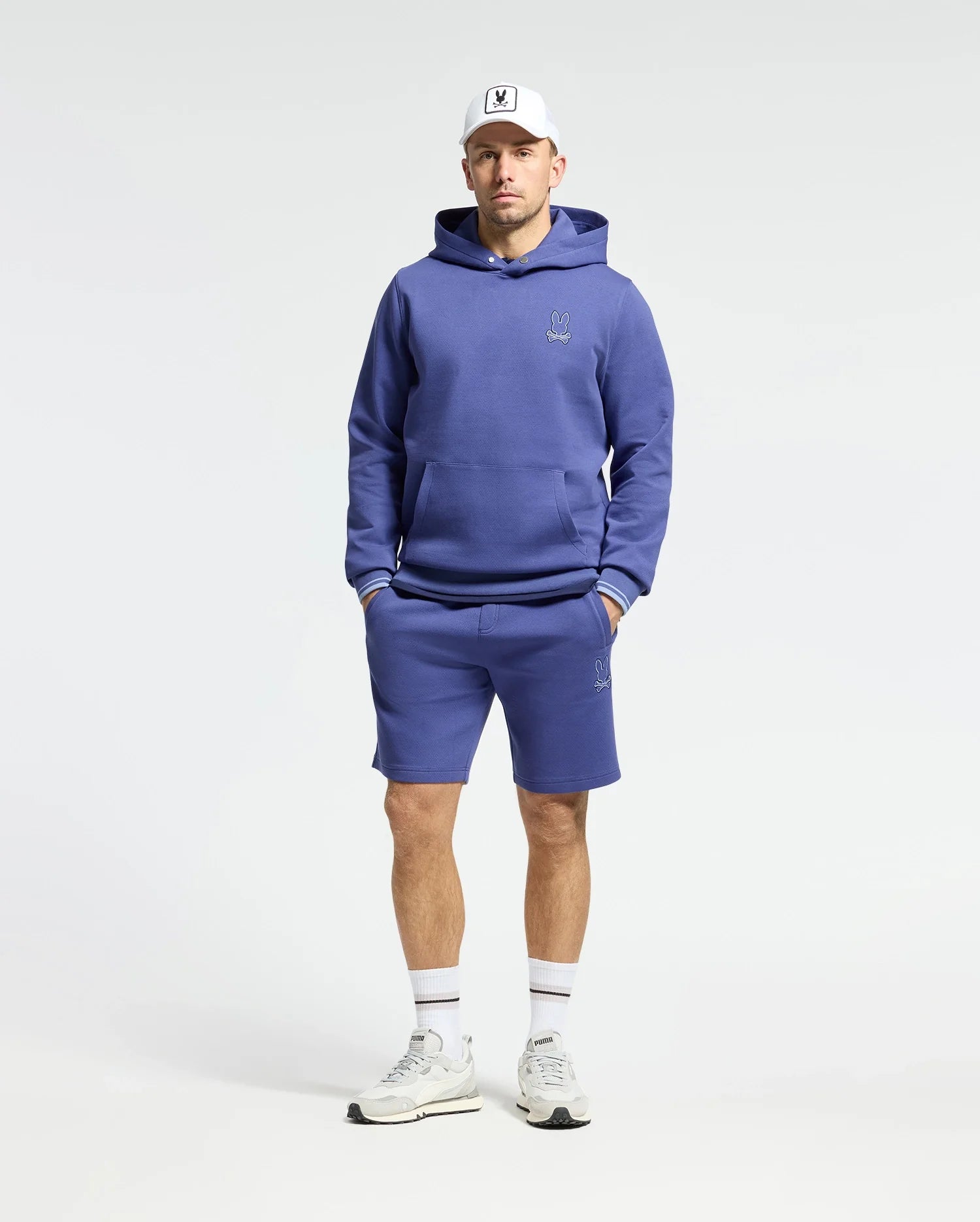 A man models the Psycho Bunny MENS DACANO PERFORATED HOODIE in purple, crafted from a stretch cotton-blend jersey with matching shorts featuring an embroidered logo. He completes the look with white sneakers and a cap, his hands resting in the hoodie pocket against a plain white backdrop.