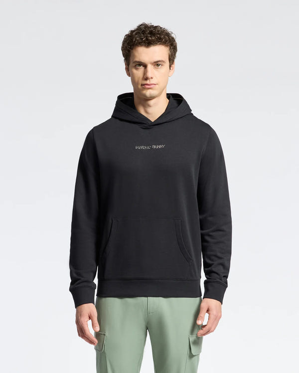 A person with curly hair wears a black Psycho Bunny Nora Classic French Terry Hoodie featuring an embroidered logo on the chest, paired with light green pants. They stand against a simple, light gray background, exuding casual comfort.