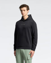 A man faces the camera in a Psycho Bunny MENS NORA CLASSIC FRENCH TERRY HOODIE - B6H434F200, featuring an embroidered chest logo, paired with light green cargo pants against a plain, light backdrop.
