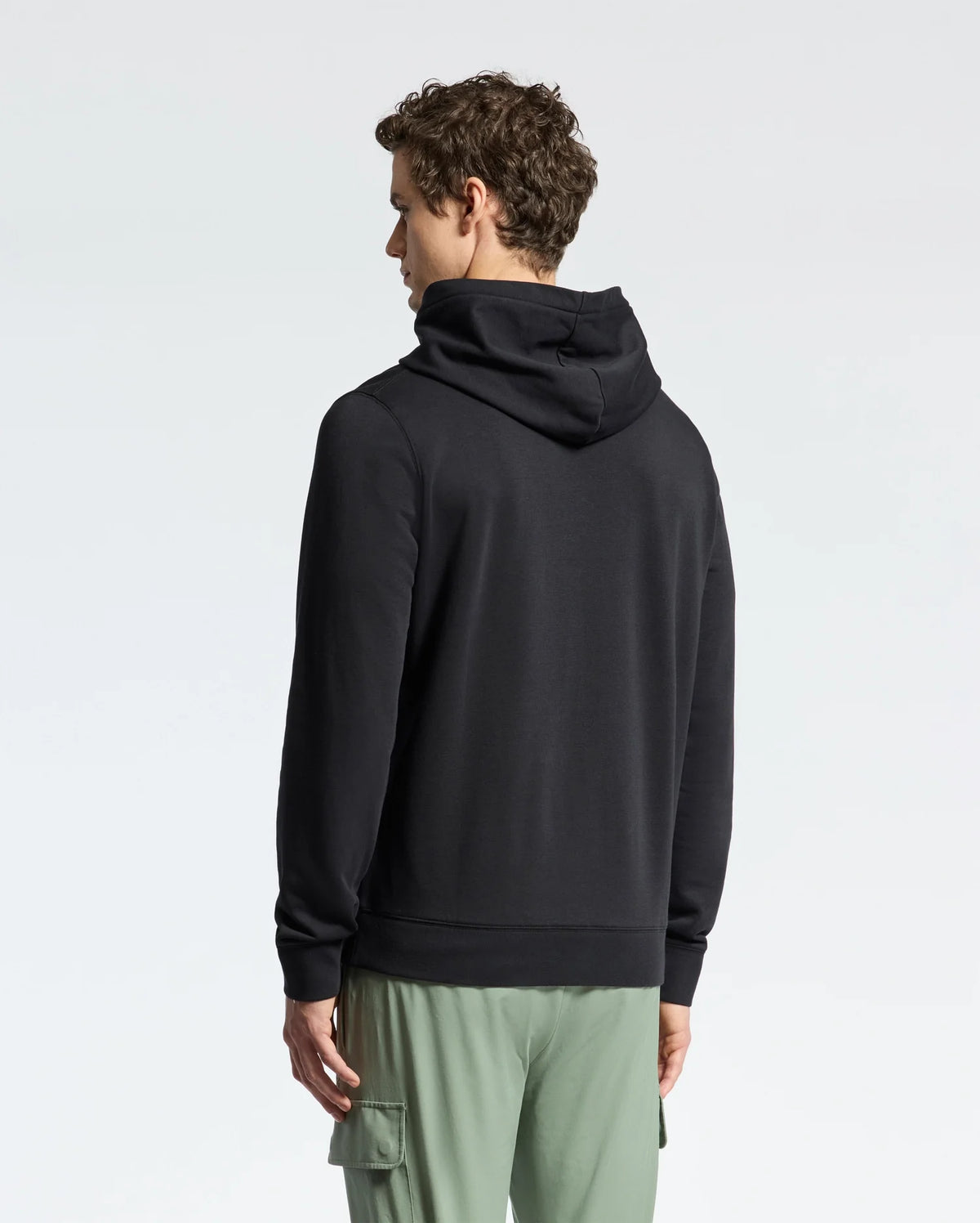 A person with curly hair is facing away, wearing a black Psycho Bunny MENS NORA CLASSIC FRENCH TERRY HOODIE and green cargo pants against a plain white background.