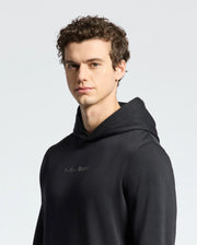 A person with curly dark hair wearing a black Mens Nora Classic French Terry Hoodie by Psycho Bunny stands against a plain light background, looking slightly to the side. The hoodie features a subtle embroidered logo across the chest.