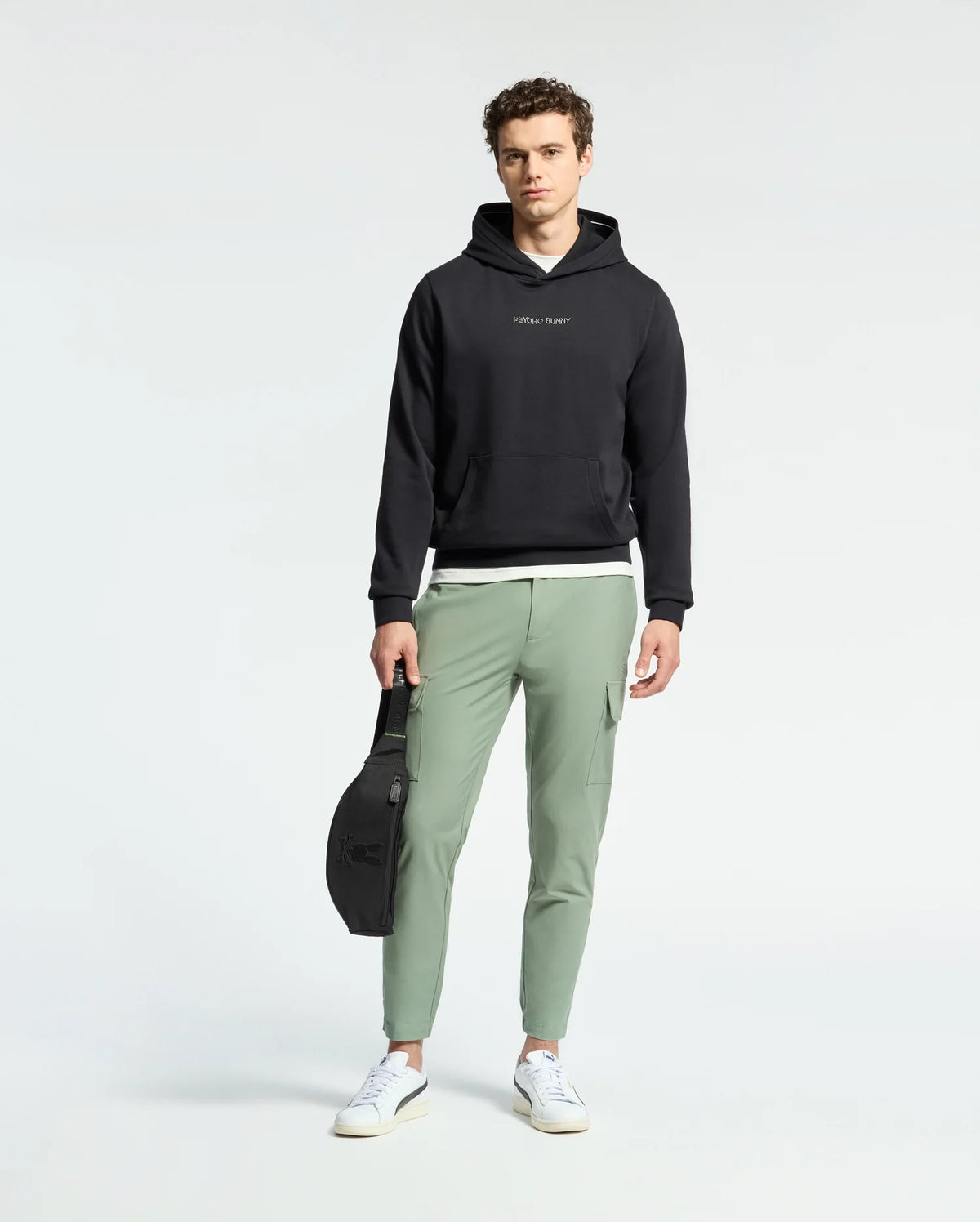 A person with curly brown hair stands against a plain background wearing light green pants, white sneakers, and a black Psycho Bunny Nora Classic French Terry Hoodie. They hold a black bag in one hand, completing their casual look.