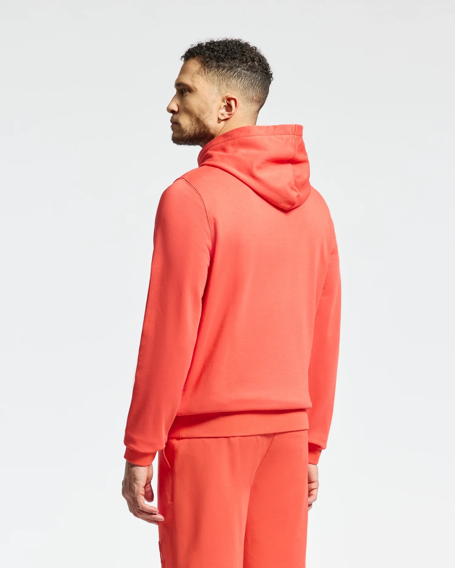 A man with short curly hair, facing left, wears a vibrant Psycho Bunny MENS NORA CLASSIC FRENCH TERRY HOODIE (B6H434F200) and matching joggers. The bright red set features an embroidered wordmark on the chest, against a plain white background.
