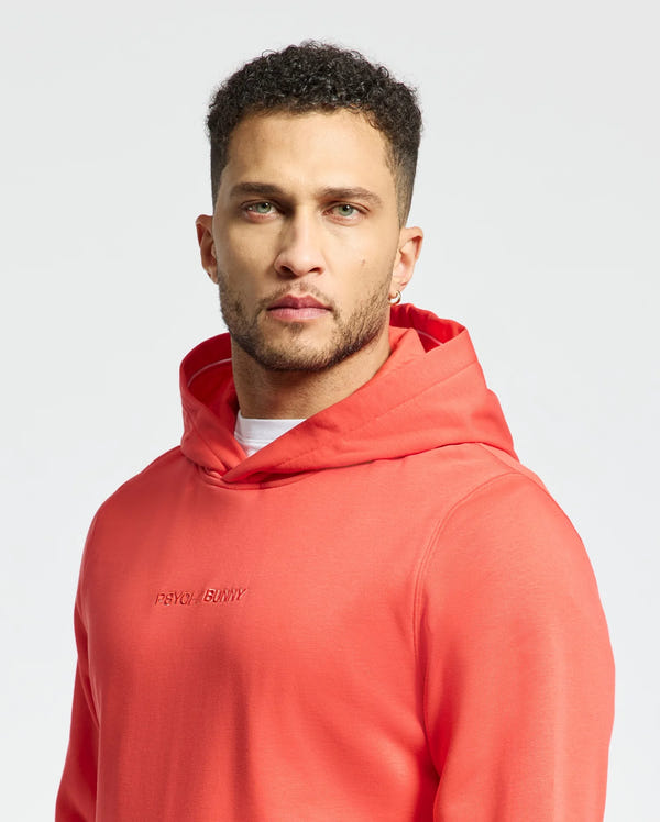 A man with short curly hair and a beard wears the coral Psycho Bunny MENS NORA CLASSIC FRENCH TERRY HOODIE - B6H434F200, showcasing an embroidered wordmark, as he stands against a plain white background.