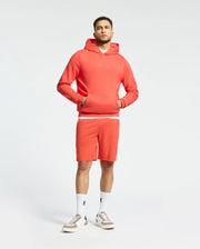A man is wearing a matching red Psycho Bunny MENS NORA CLASSIC FRENCH TERRY HOODIE and shorts, with his hands in the hoodie's front pocket. He pairs this with white socks featuring a black embroidered logo and pristine white sneakers against a light backdrop.