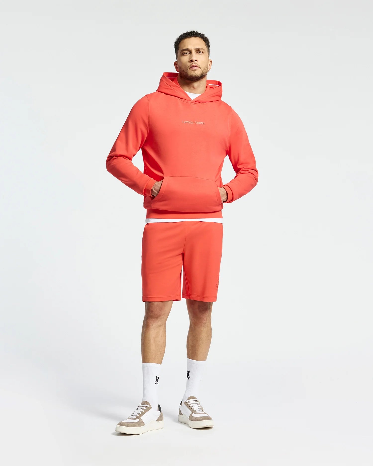 A man is wearing a matching red Psycho Bunny MENS NORA CLASSIC FRENCH TERRY HOODIE and shorts, with his hands in the hoodie's front pocket. He pairs this with white socks featuring a black embroidered logo and pristine white sneakers against a light backdrop.