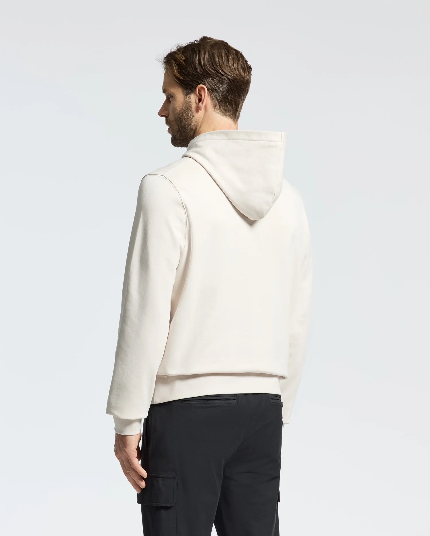 A man with short hair, seen from the side and slightly behind, wears a Psycho Bunny MENS NORA CLASSIC FRENCH TERRY HOODIE—B6H434F200. The light-colored design features a subtle embroidered logo. He pairs it with black pants against a plain white background, while the hood lays flat on his back.
