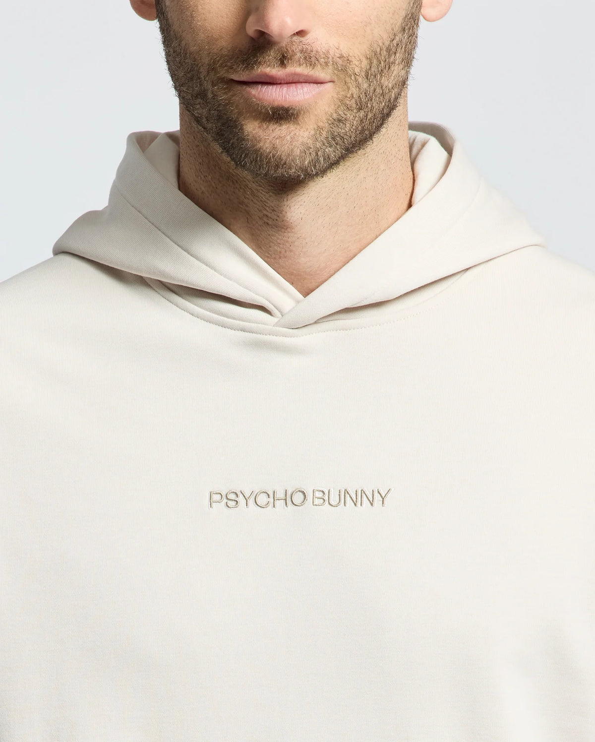 A bearded man is wearing a cream MENS NORA CLASSIC FRENCH TERRY HOODIE by Psycho Bunny with "PSYCHO BUNNY" embroidered on the front against a plain white background.