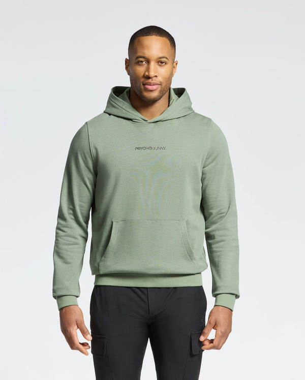 A man poses against a white backdrop in a Psycho Bunny light green MENS NORA CLASSIC FRENCH TERRY HOODIE - B6H434F200 with an embroidered logo. He pairs it with black pants, his short hair enhancing the simple scene.