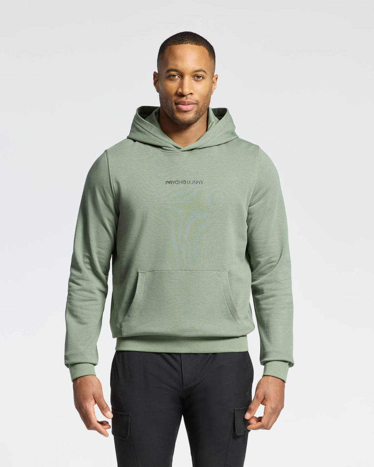 A man poses against a white backdrop in a Psycho Bunny light green MENS NORA CLASSIC FRENCH TERRY HOODIE - B6H434F200 with an embroidered logo. He pairs it with black pants, his short hair enhancing the simple scene.