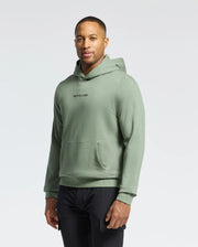 A person in a green Psycho Bunny Men's Nora Classic French Terry Hoodie (B6H434F200) and black pants stands against a plain white background. They have short hair, a relaxed expression, and their hands at their sides, showcasing the embroidered wordmark logo on the hoodie.