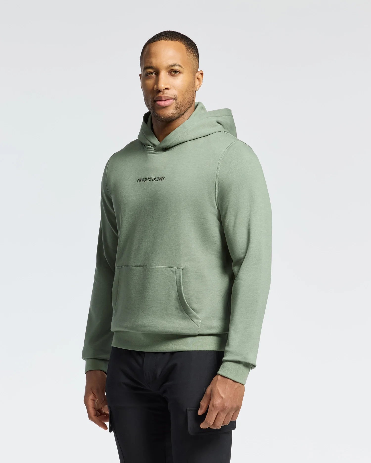 A person in a green Psycho Bunny Men's Nora Classic French Terry Hoodie (B6H434F200) and black pants stands against a plain white background. They have short hair, a relaxed expression, and their hands at their sides, showcasing the embroidered wordmark logo on the hoodie.