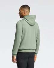 A man in the Psycho Bunny MENS NORA CLASSIC FRENCH TERRY HOODIE - B6H434F200, featuring an embroidered logo, stands back to camera against a white background. His cozy green hoodie contrasts sharply with his black pants.