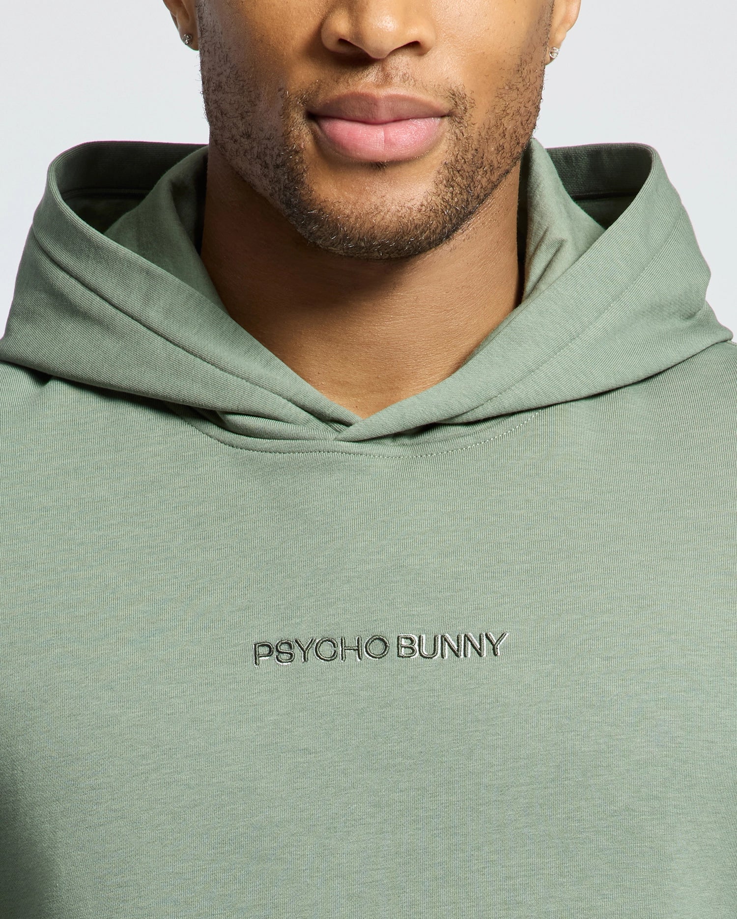 A person wears a light green Psycho Bunny Nora Hoodie, crafted from French terry fabric. The focus is on the upper body, highlighting the distinctive "Psycho Bunny" wordmark logo embroidered on the front.