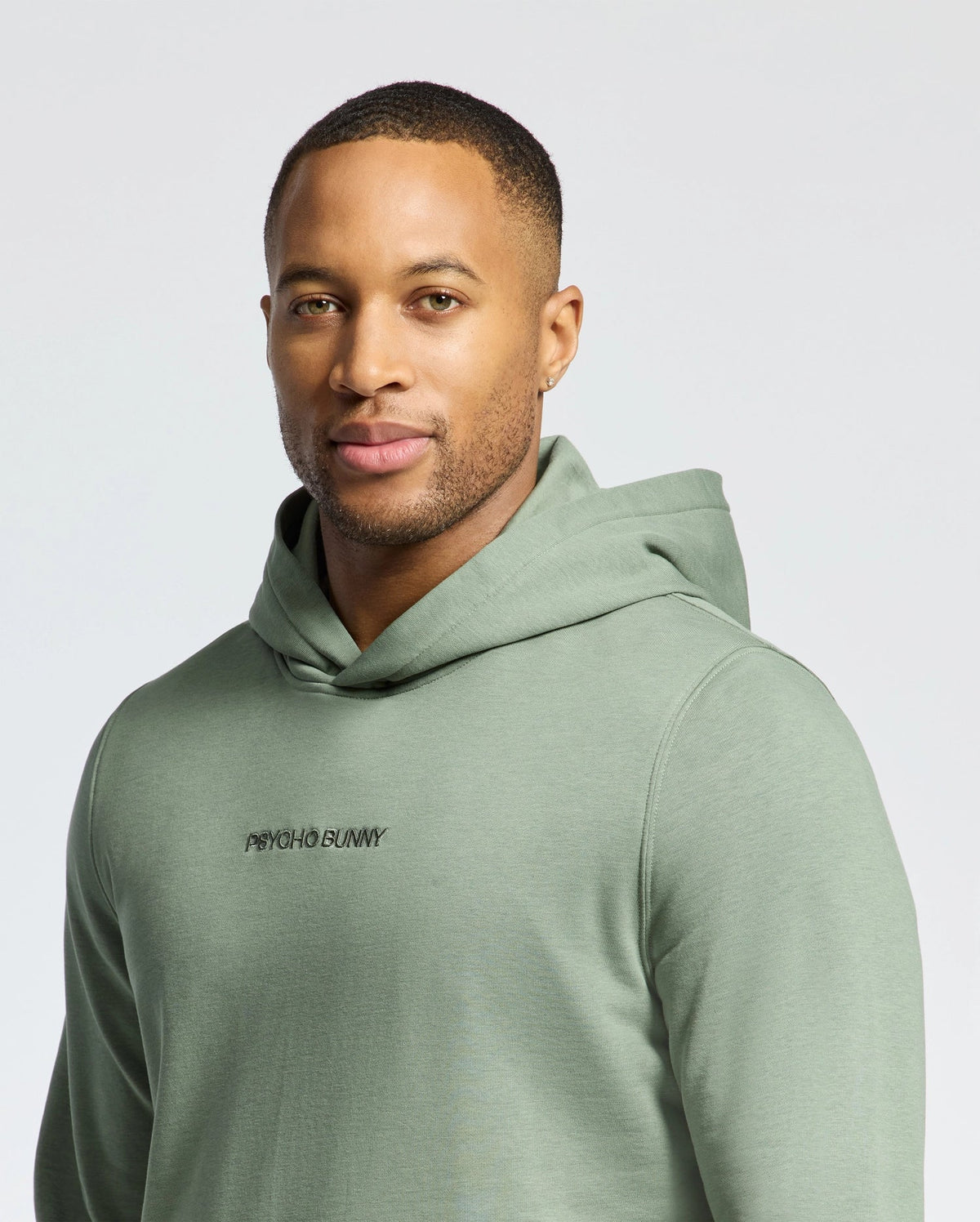 A person is wearing a light green MENS NORA CLASSIC FRENCH TERRY HOODIE by Psycho Bunny, featuring the embroidered wordmark logo. The relaxed fit and design exude calmness as they're seen against a neutral background.