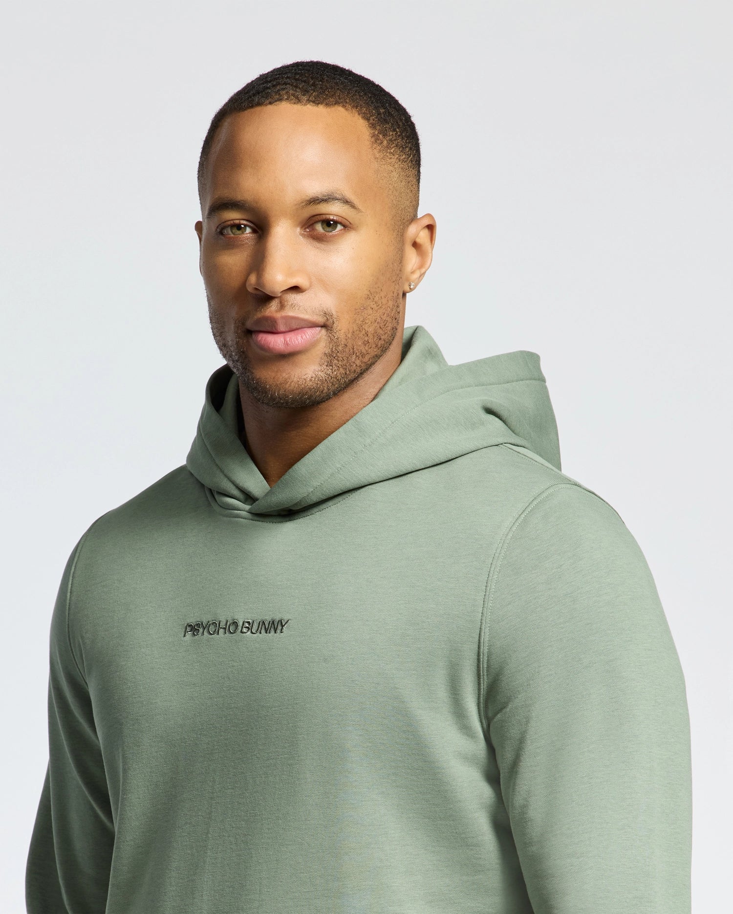 A person is wearing a light green MENS NORA CLASSIC FRENCH TERRY HOODIE by Psycho Bunny, featuring the embroidered wordmark logo. The relaxed fit and design exude calmness as they're seen against a neutral background.