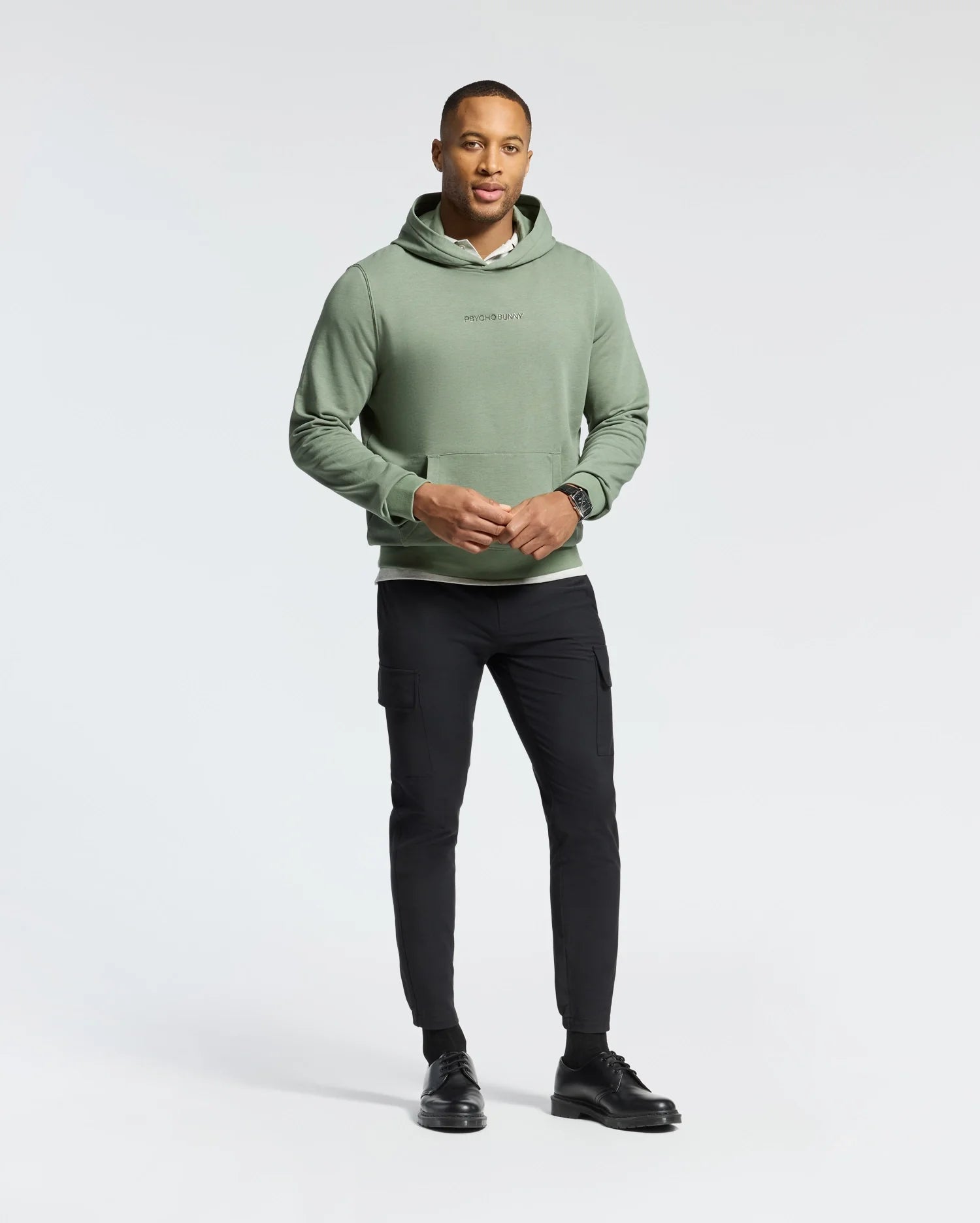A man stands against a plain background wearing a Psycho Bunny MENS NORA CLASSIC FRENCH TERRY HOODIE in light green. Its soft fabric and embroidered wordmark logo offer understated style. He pairs it with black pants and shoes, gazing at the camera with his hands together.