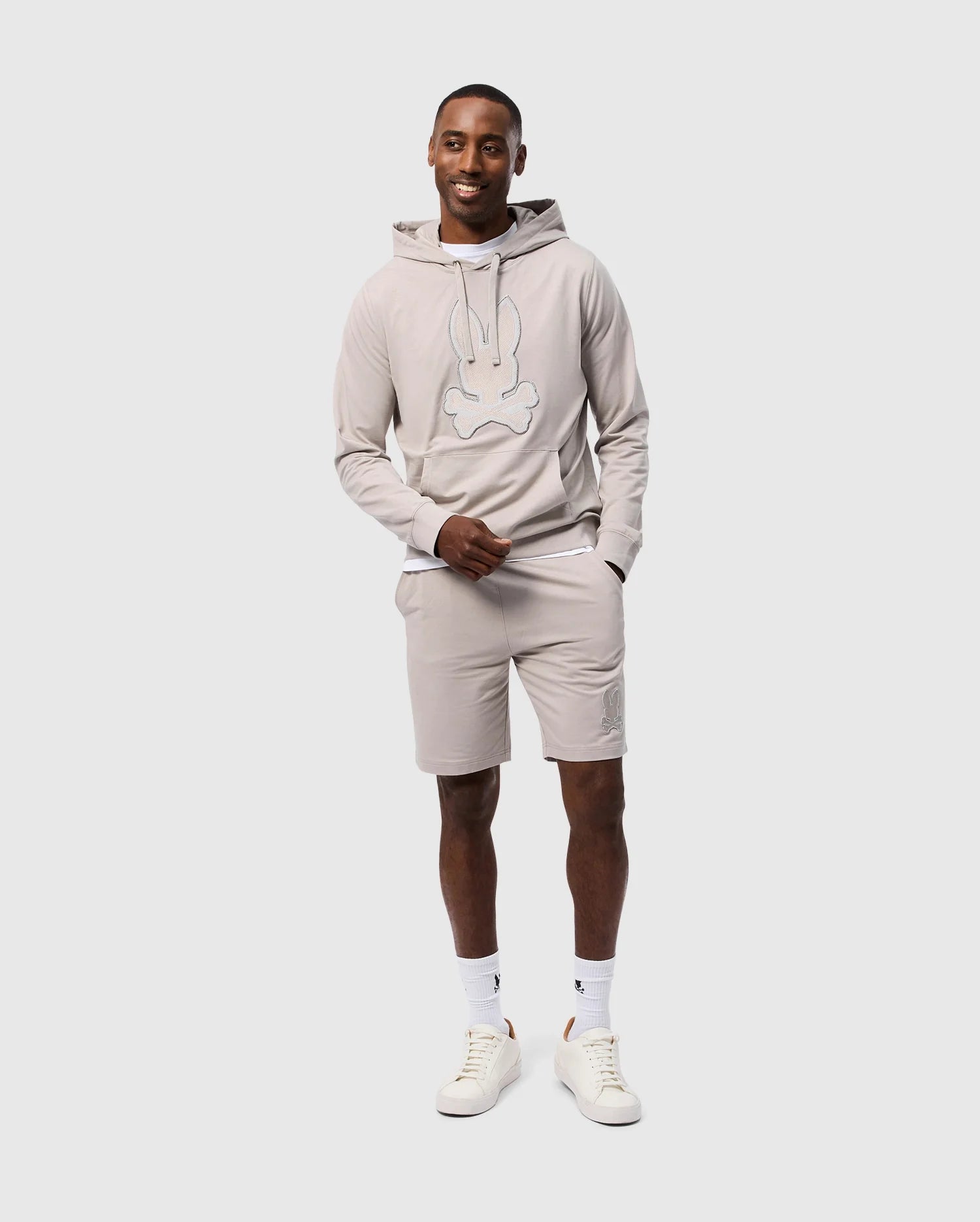 A man stands smiling, dressed in a matching beige MENS WALTER LIGHTWEIGHT HOODIE - B6H476D200 and shorts set by Psycho Bunny, crafted from Micro French terry. His regular fit hoodie features a large, stylized cartoon bunny face. He is wearing white sneakers with beige accents and white socks. One hand rests in his shorts pocket as he stands against a plain white background.