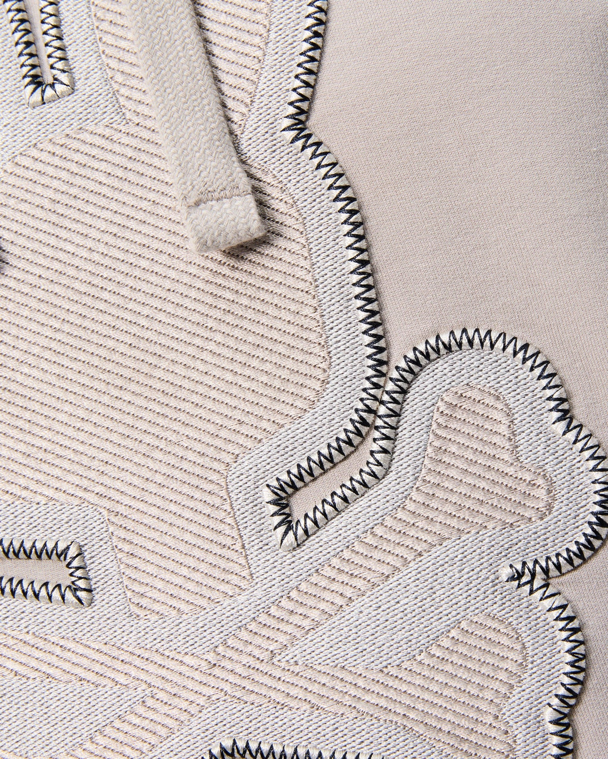 Close-up of the textured fabric displaying embroidered details. The material is beige Micro French terry with raised, woven patches in a slightly lighter tone bordered by black zigzag stitches. A glimpse of the beige drawstring, typical of the regular fit "MENS WALTER LIGHTWEIGHT HOODIE - B6H476D200" from Psycho Bunny, is visible at the top.