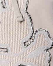 Close-up of the textured fabric displaying embroidered details. The material is beige Micro French terry with raised, woven patches in a slightly lighter tone bordered by black zigzag stitches. A glimpse of the beige drawstring, typical of the regular fit "MENS WALTER LIGHTWEIGHT HOODIE - B6H476D200" from Psycho Bunny, is visible at the top.