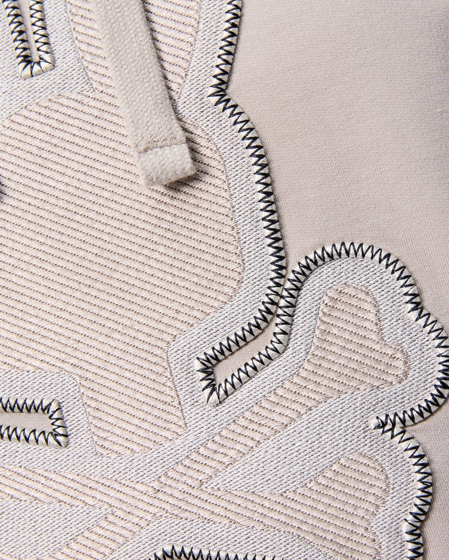 Close-up of the textured fabric displaying embroidered details. The material is beige Micro French terry with raised, woven patches in a slightly lighter tone bordered by black zigzag stitches. A glimpse of the beige drawstring, typical of the regular fit "MENS WALTER LIGHTWEIGHT HOODIE - B6H476D200" from Psycho Bunny, is visible at the top.