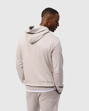 A person wearing a Psycho Bunny MENS WALTER LIGHTWEIGHT HOODIE - B6H476D200 in regular fit beige with the hood down faces away from the camera. The Micro French terry hoodie has "PSYCHO BUNNY" embroidered near the bottom. The person has one hand in their pocket and is dressed in matching beige pants against a plain background.