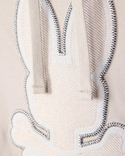 Close-up view of an embroidered bunny outline on beige Micro French terry fabric from the Psycho Bunny MENS WALTER LIGHTWEIGHT HOODIE - B6H476D200. The bunny is stitched with white thread and black accents, and has two strings hanging from the top, likely part of the hoodie. The texture and stitching details are clearly visible on this regular fit piece.