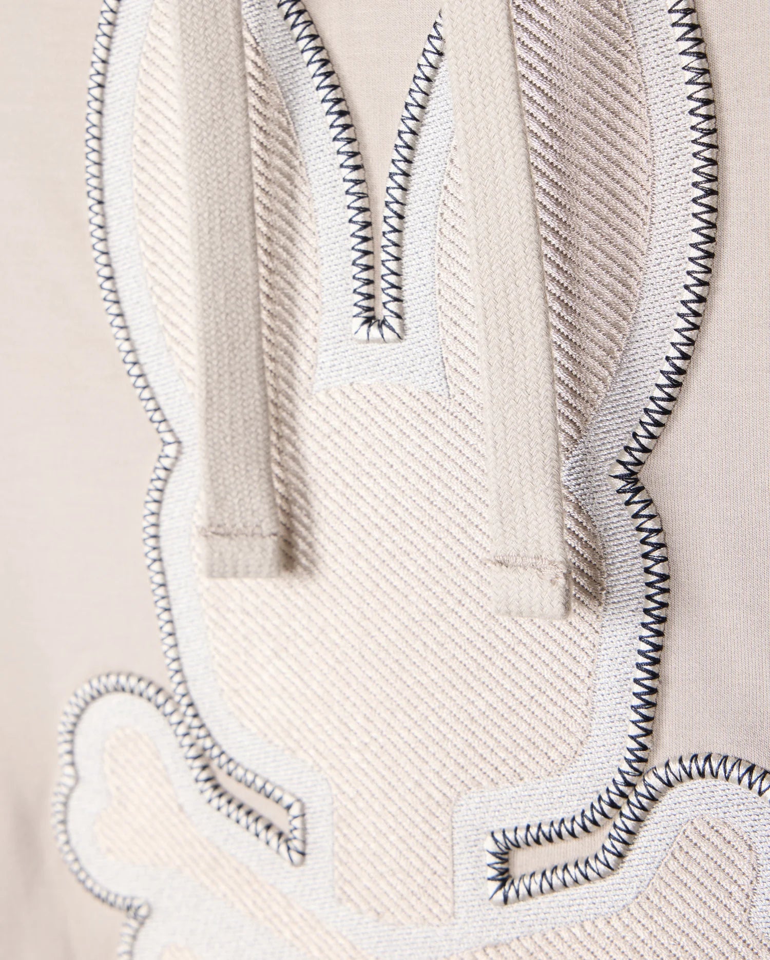 Close-up view of an embroidered bunny outline on beige Micro French terry fabric from the Psycho Bunny MENS WALTER LIGHTWEIGHT HOODIE - B6H476D200. The bunny is stitched with white thread and black accents, and has two strings hanging from the top, likely part of the hoodie. The texture and stitching details are clearly visible on this regular fit piece.
