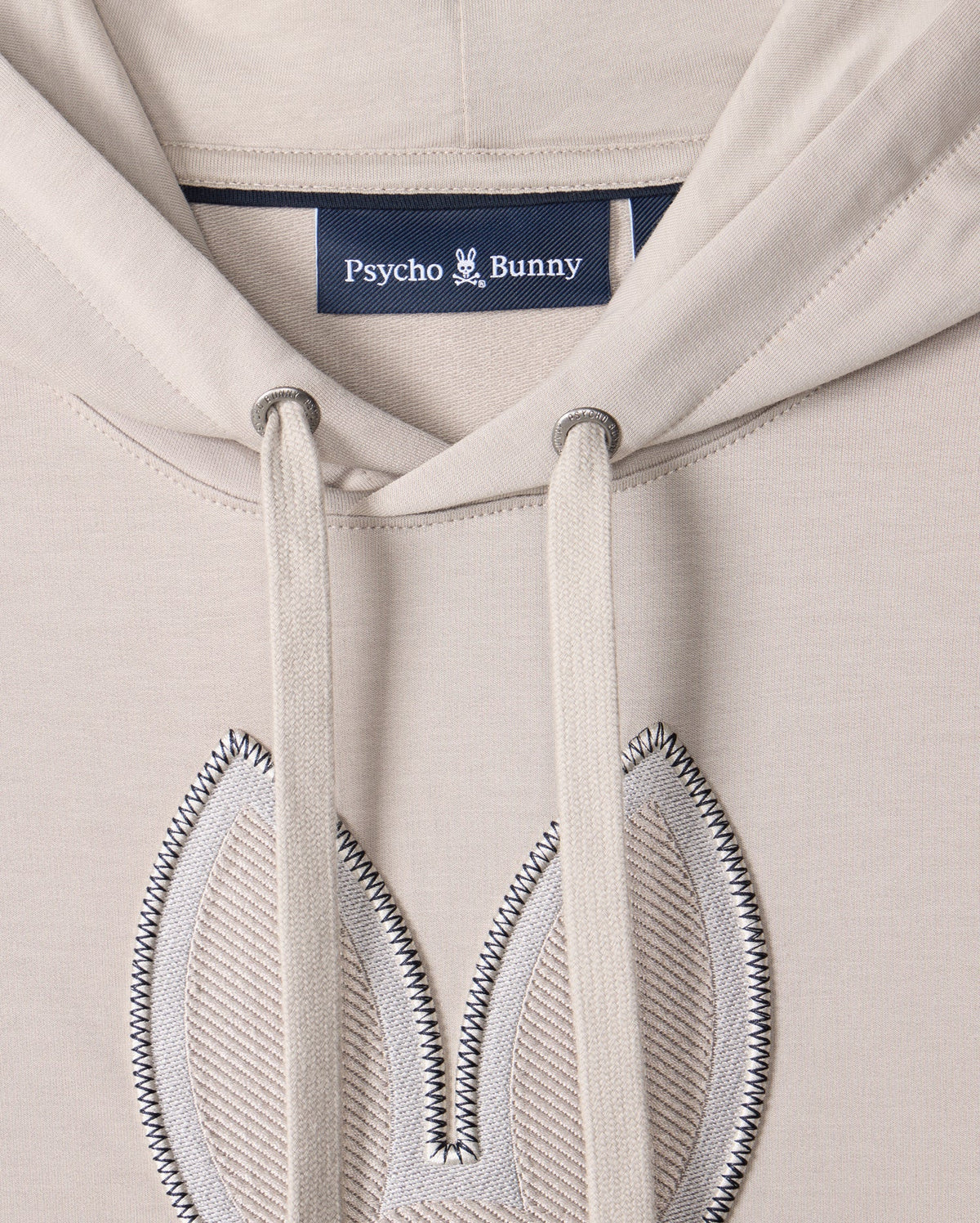 Close-up of the MENS WALTER LIGHTWEIGHT HOODIE - B6H476D200 by Psycho Bunny, crafted from beige Micro French terry. This regular fit hoodie features two drawstrings and an embroidered bunny ear design on the chest area, with the "Psycho Bunny" label sewn inside.